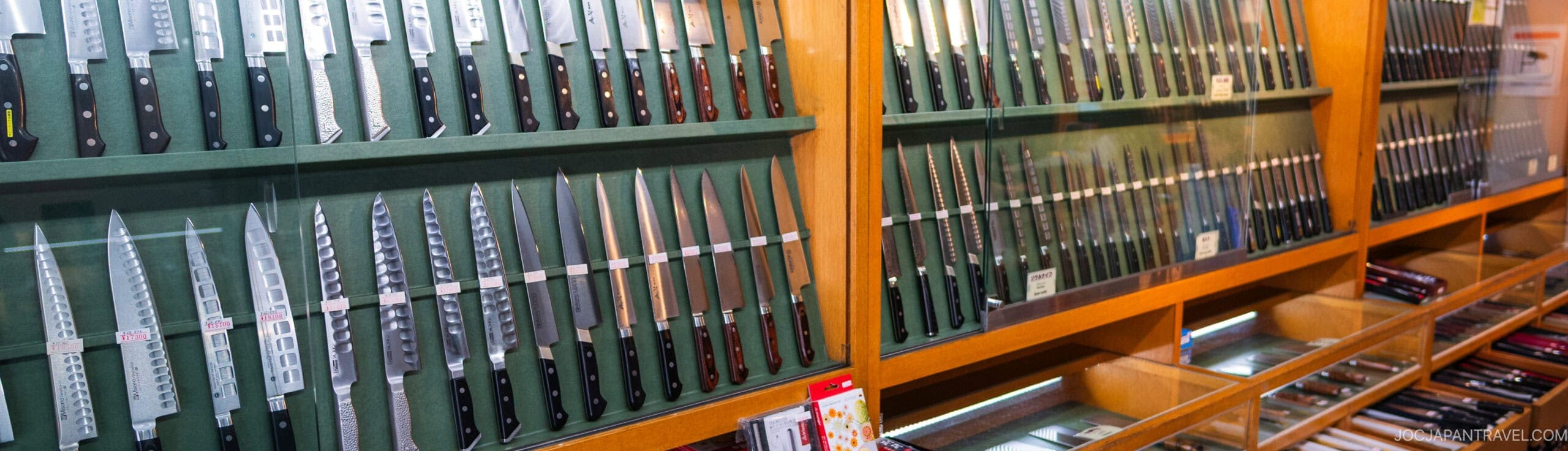 A display case filled with various kitchen knives arranged neatly in rows, showing different blade shapes and handle styles, with additional knives stored in drawers below.