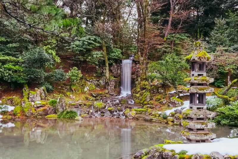 A tranquil Japanese garden, inspired by Kenrokuen Garden, features a small waterfall flowing into a pond, surrounded by lush greenery and moss-covered rocks. A stone lantern stands by the water, with patches of snow scattered around.