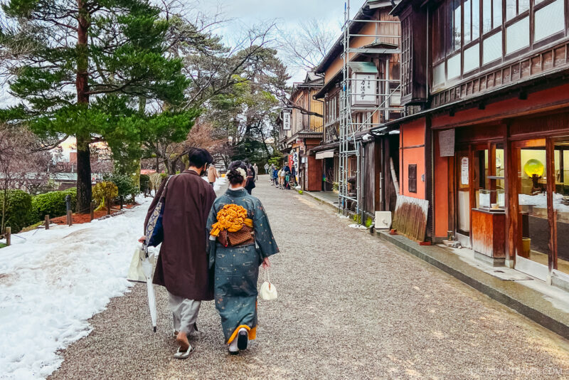 A couple wearing traditional Japanese clothing walks down a quiet street lined with wooden buildings and shops near Kenrokuen Garden; snow rests on the ground near trees.
