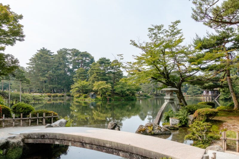 A serene Japanese garden inspired by Kenrokuen Garden, featuring a stone bridge over a calm pond, lush green trees, shrubs, and traditional stone lanterns that reflect a peaceful, natural landscape.