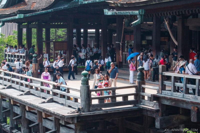 A diverse group of people, including tourists with cameras and some in traditional Japanese clothing, stand on the wooden terrace of Kiyomizu-dera temple with ornate details and a lush backdrop.