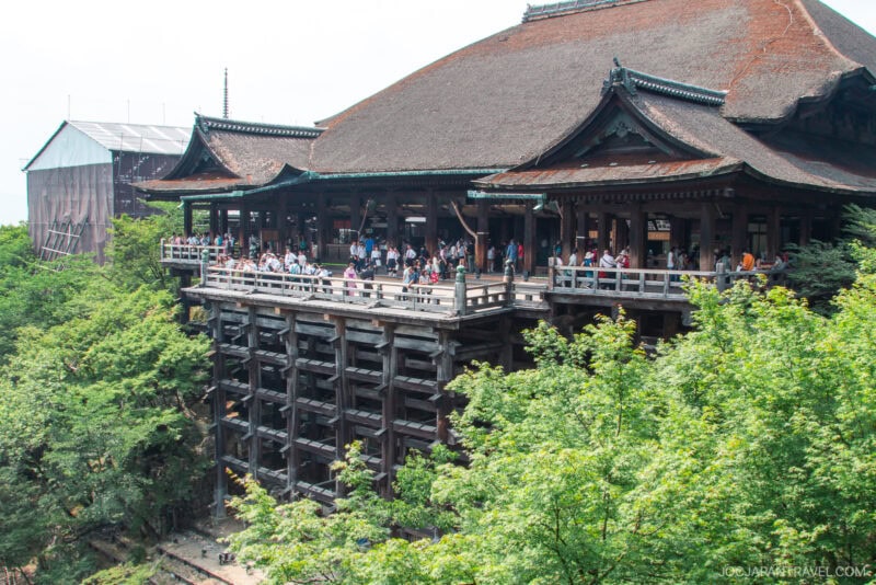 A large wooden temple, Kiyomizu-dera, with a wide veranda filled with visitors, overlooks green trees. The building features traditional Japanese architectural elements and sits elevated on tall wooden pillars.