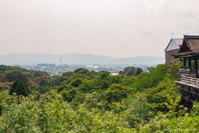 A view from the wooden terrace of Kiyomizu-dera captures a lush green forest, distant city buildings, and hazy mountains under a muted sky.