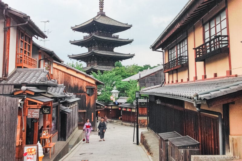 A narrow street in Kyoto, Japan, lined with traditional wooden buildings. Two people in yukatas stroll toward a tall pagoda near Kiyomizu-dera, surrounded by greenery. The scene feels both peaceful and historic.
