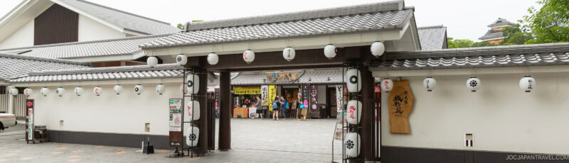 Traditional Japanese building entrance with white walls, tiled roof, and hanging paper lanterns; people are gathered inside near colorful banners, and a historic castle is visible in the background.