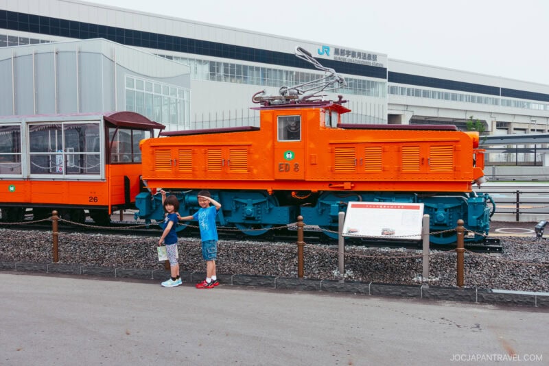 Two boys stand and pose in front of a bright orange electric locomotive displayed outdoors at a railway museum near Kurobe Gorge; a modern building and another train car are visible in the background.