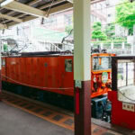 Kurobe Gorge (黒部峡谷): Japan’s Natural Wonder by Scenic Torokko Train Bright orange train cars at a station platform in Japans scenic Kurobe Gorge, with greenery and buildings in the background. The train has an old-fashioned design and is partially covered by a roof.