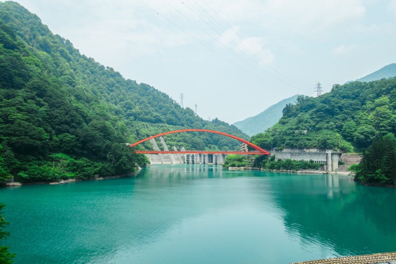 A bright red arch bridge spans the calm turquoise river of Kurobe Gorge, surrounded by lush green mountains under a partly cloudy sky. A concrete dam structure is visible beneath the bridge.