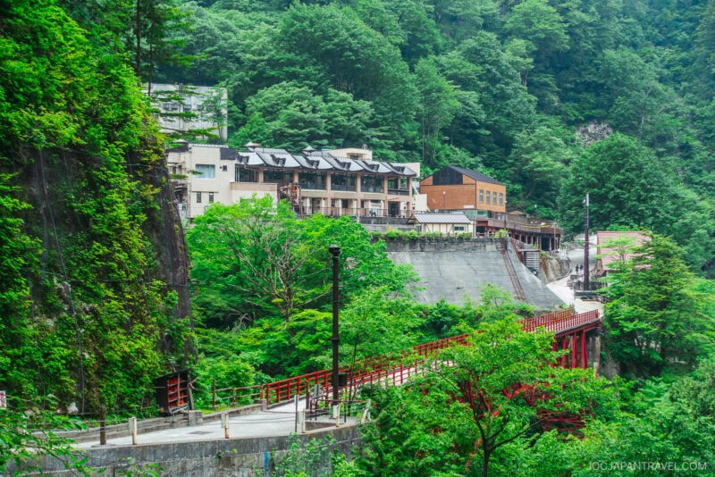 A cluster of buildings and a red bridge nestle among lush green trees on a steep hillside in the mountainous Kurobe Gorge, creating a serene and picturesque scene.