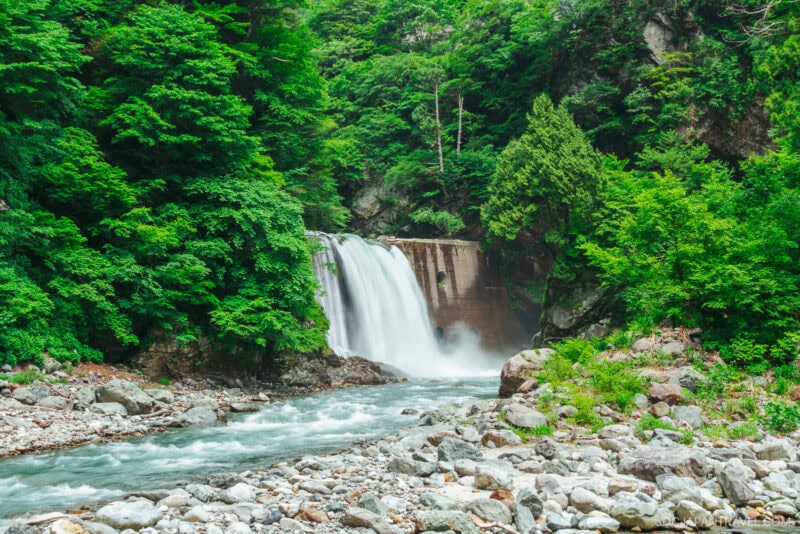 A waterfall cascades over a concrete barrier into a rocky river in Kurobe Gorge, surrounded by lush, dense green forest and trees. Rocks line the riverbank in the foreground.