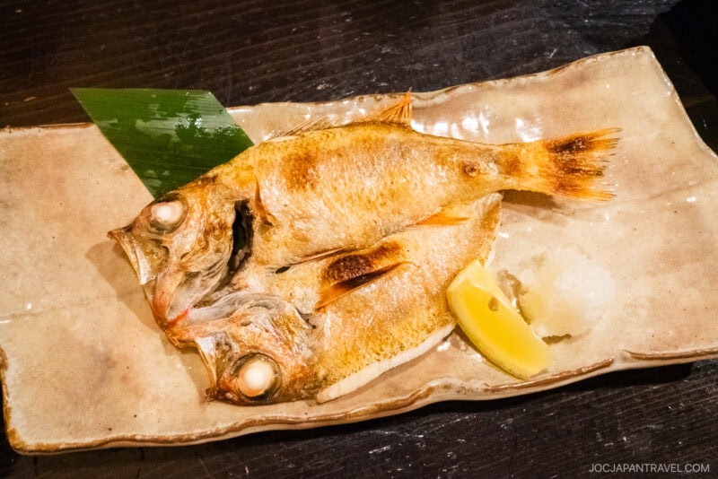 At Kuroya Izakaya Kanazawa, enjoy two whole grilled fish with crispy skin, served on a rectangular ceramic plate with a wedge of lemon, radish garnish, and a green leaf on a dark wooden table.