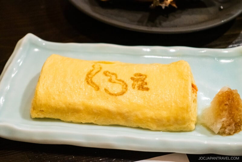 A rectangular Japanese omelette (tamagoyaki) with a brown design on top is served on a light blue plate at Kuroya Izakaya Kanazawa, accompanied by a mound of grated daikon radish on the side.