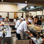 Kuroyuri (黒百合): Delicious Oden in Kanazawa Station Chefs in white uniforms prepare food behind the sushi counter at Kuroyuri Oden Kanazawa, while customers sit around, eating and watching. Bottles and dishes line the counter, creating a lively atmosphere in this Japanese restaurant.