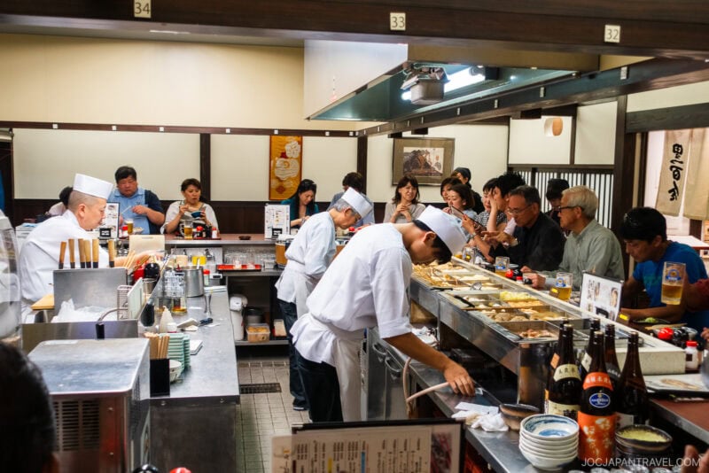 Chefs in white uniforms prepare food behind the sushi counter at Kuroyuri Oden Kanazawa, while customers sit around, eating and watching. Bottles and dishes line the counter, creating a lively atmosphere in this Japanese restaurant.