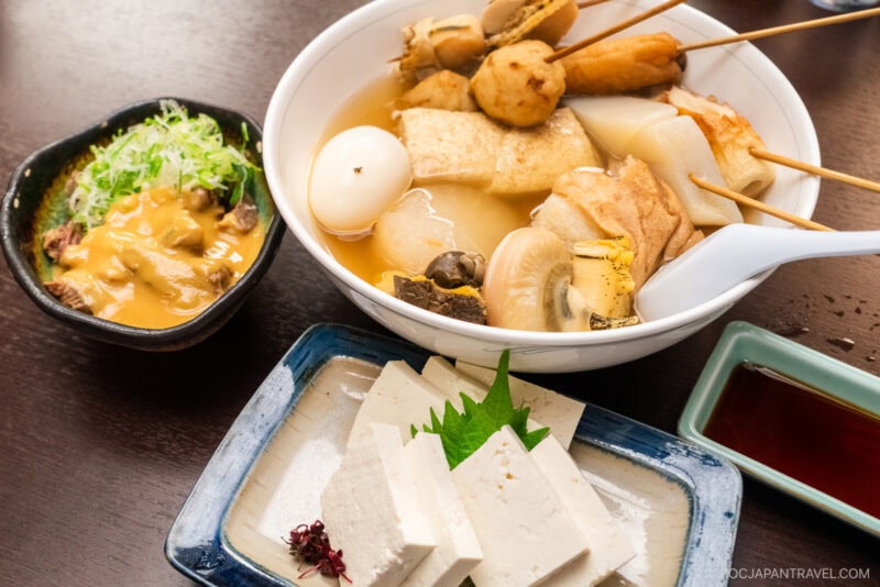 A bowl of Kuroyuri Oden Kanazawa with assorted skewered ingredients, a dish of fresh tofu slices with garnish, a small bowl of miso sauce with green onions, and a soy sauce dish on a wooden table.