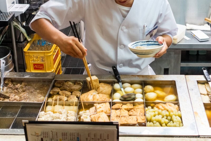 At Kuroyuri Oden Kanazawa, a person in a white chef’s coat serves Japanese oden from a steaming counter filled with simmering eggs, tofu, and vegetables, using chopsticks and a ladle.