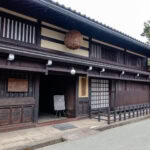 Kusakabe Heritage House (日下部民藝館) Traditional Japanese wooden building with dark timber exterior, sliding doors, and a large round brown sugidama above the entrance—this serene Kusakabe Heritage House sits on a quiet street lined with trees in the background.