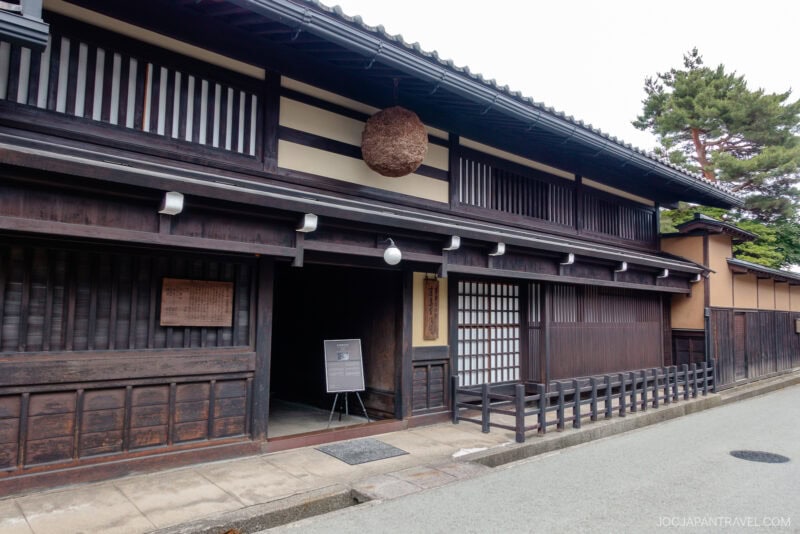 Traditional Japanese wooden building with dark timber exterior, sliding doors, and a large round brown sugidama above the entrance&mdash;this serene Kusakabe Heritage House sits on a quiet street lined with trees in the background.