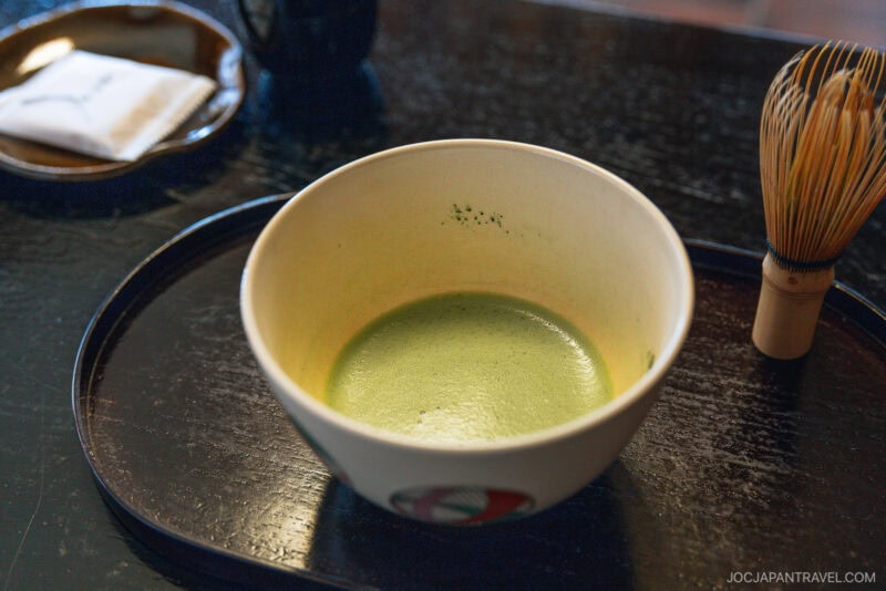 A bowl of frothy matcha green tea sits on a black tray, next to a bamboo whisk and a plate with a folded napkin—an essential experience for those exploring Kyoto what to eat.