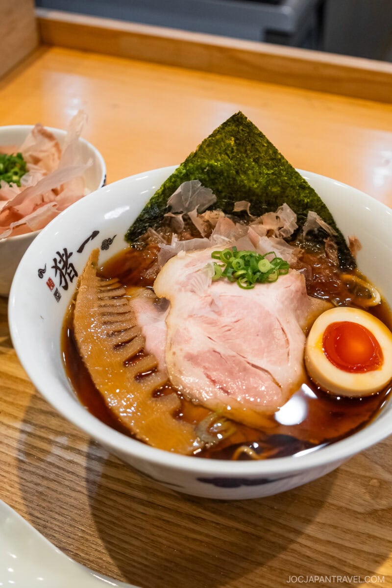 A bowl of ramen with sliced pork, half a soft-boiled egg, green onions, bamboo shoots, seaweed, and bonito flakes in a rich, dark broth—Kyoto what to eat for an authentic taste—served on a wooden counter.