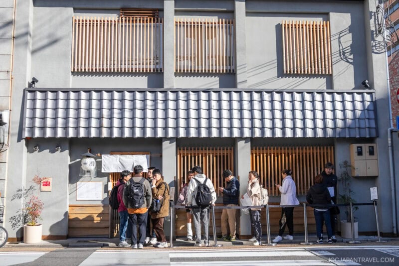 A group of people stands in line outside a modern Japanese-style restaurant with wooden slats and a tiled roof on a sunny day, perfect for anyone curious about Kyoto what to eat.