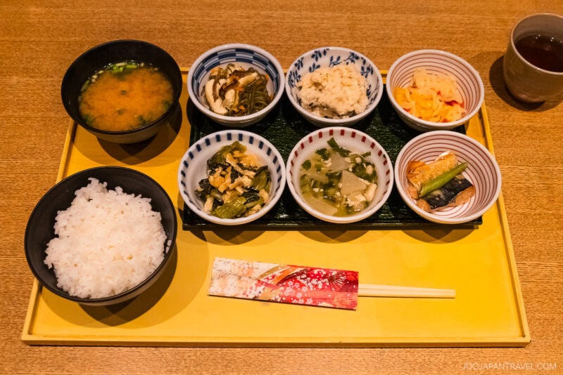 A Japanese meal set on a yellow tray with white rice, miso soup, six small dishes of vegetables, tofu, pickles, tea, and decorated chopsticks—perfect for those wondering in Kyoto what to eat for an authentic experience.