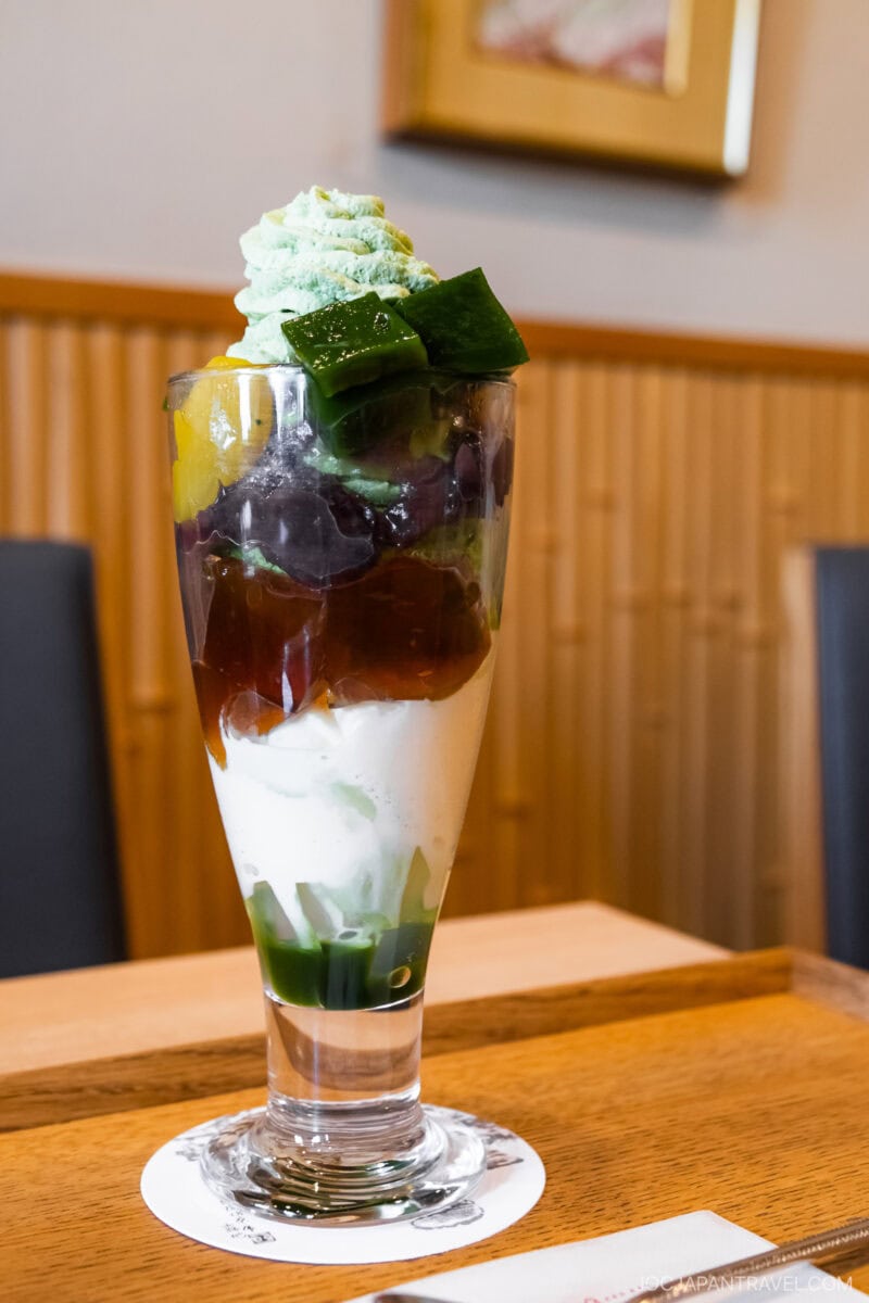 A tall parfait glass filled with layers of green jelly, whipped cream, red beans, and topped with matcha whipped cream and green jelly cubes—this dessert is a must-try in Kyoto if you’re wondering what to eat in the city.