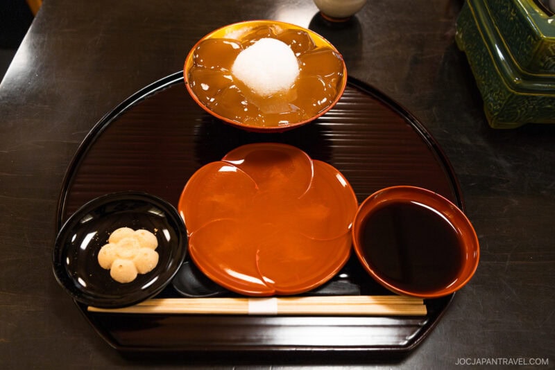 A traditional Japanese dessert set on a lacquered tray, featuring warabi mochi with syrup and a topping, is a must-try in Kyoto. Wondering what to eat? Enjoy round sweets, rich dark syrup, and wooden chopsticks for an authentic experience.