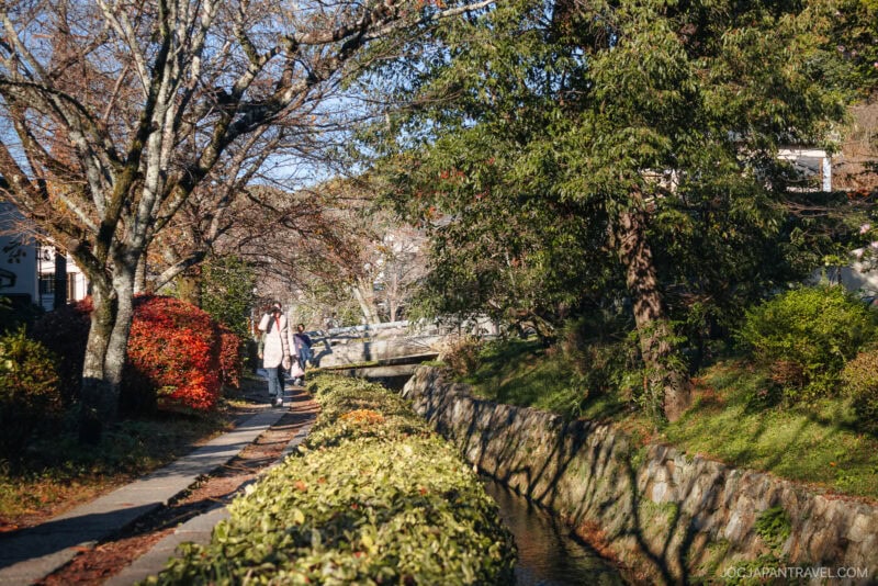 A narrow stone path—reminiscent of the Philosopher’s Path—runs alongside a small canal bordered by trees in autumn colors. Two people walk under dappled sunlight, while a small bridge crosses the canal in the background.