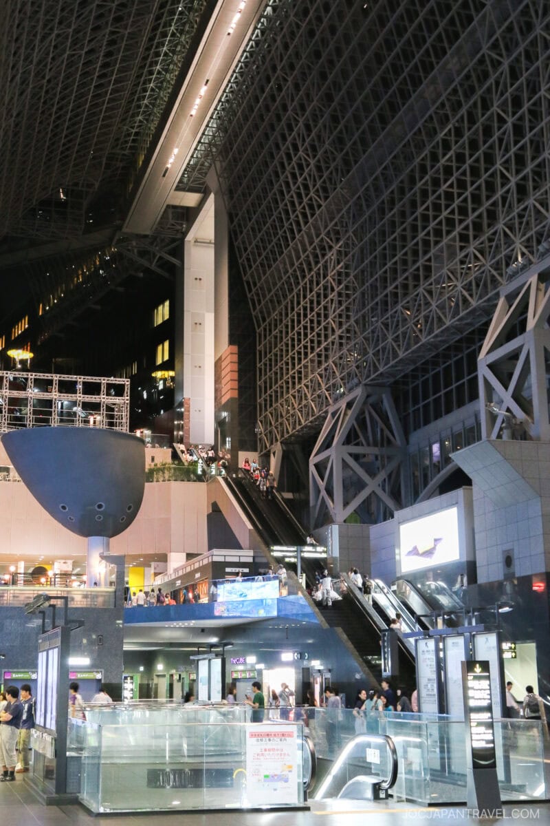 A spacious, modern Kyoto Station interior with high glass ceilings, multiple floors, bright lighting, escalators, and people walking or waiting. Digital screens and structural steel beams are featured throughout this bustling hub.