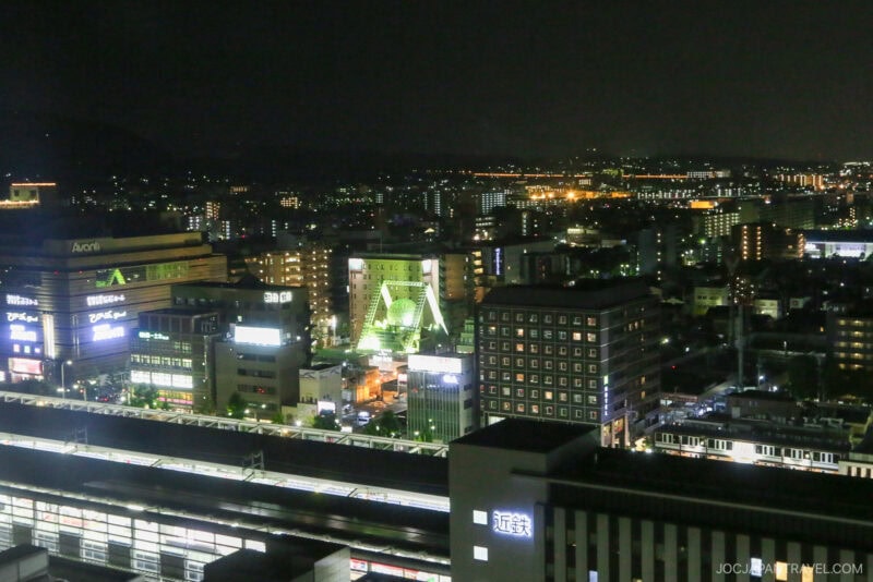 A nighttime cityscape featuring illuminated buildings, bustling streets, and the well-lit Kyoto Station in the foreground, with distant city lights stretching across the horizon.