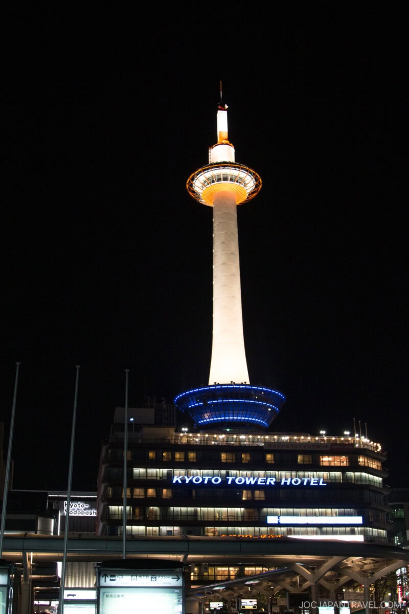 Kyoto Tower illuminated at night, glowing orange and white above the Kyoto Tower Hotel building with signs visible, stands tall near Kyoto Station against a dark sky.