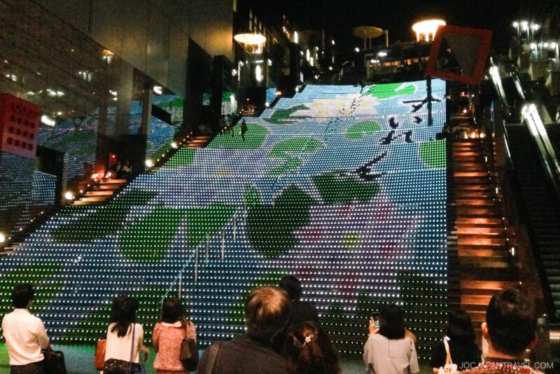 People stand at the base of a large, wide staircase at Kyoto Station at night, watching colorful LED lights form an illuminated mural across the steps. The scene is lively, with reflected lights and a vibrant city atmosphere.