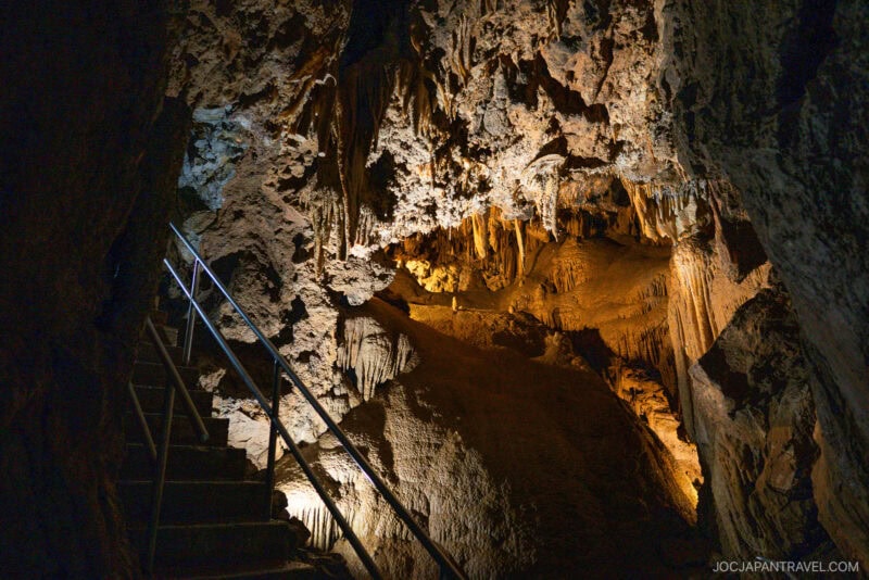 A dimly lit cave interior at Lake Shasta Caverns features rough, textured rock formations and stalactites hanging from the ceiling. A metal handrail lines steep stairs ascending along the left side, with warm lights highlighting each unique feature.