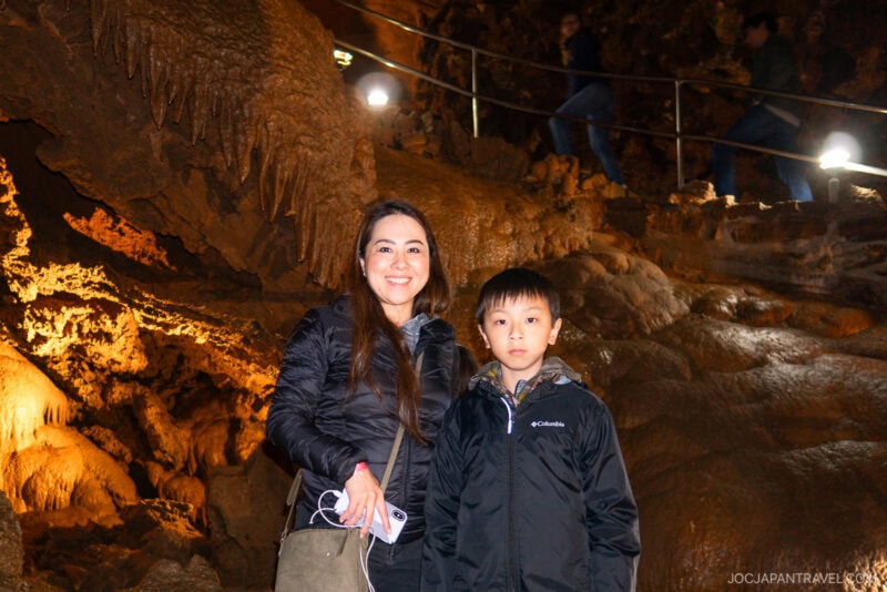 A woman and a boy, both wearing jackets, stand inside Lake Shasta Caverns among orange and brown rock formations. The boy looks serious while the woman smiles, with other visitors visible in the background on an elevated path.