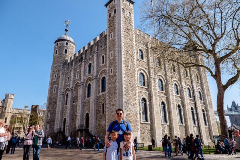 A man and two children pose in front of the historic Tower of London on a sunny day, with other visitors and a large stone building highlighting this iconic London landmark in the background.