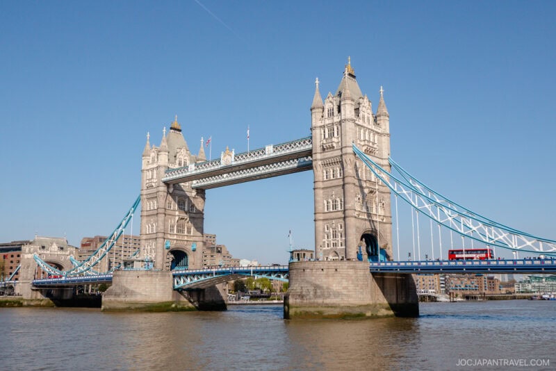 Tower Bridge in London spans the River Thames under a clear blue sky. A red double-decker bus crosses the lower level, with historic buildings and modern London structures visible in the background.
