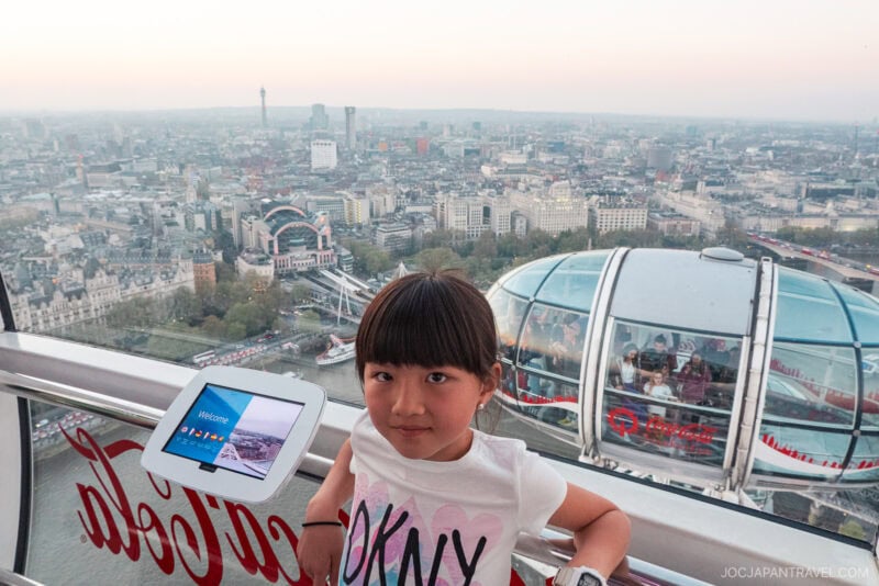 A young girl stands inside a glass capsule on the London Eye, overlooking the London skyline with various buildings visible in the background. A tablet display and reflections of the capsule’s structure are also seen.