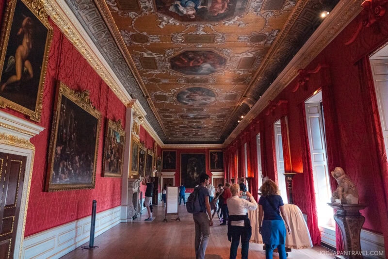Tourists admire artwork in a grand London gallery with red wallpaper, large paintings in ornate gold frames, tall windows with red curtains, and an elaborate painted ceiling. A marble statue stands near the windows.