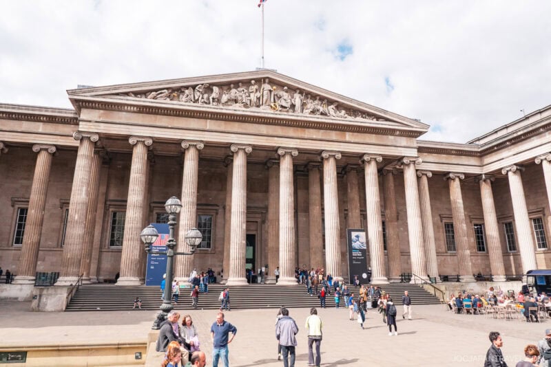 The photo shows the British Museum’s grand entrance in London, featuring tall columns and stone steps. Visitors are gathered in front, some sitting and some walking, under a partly cloudy sky.