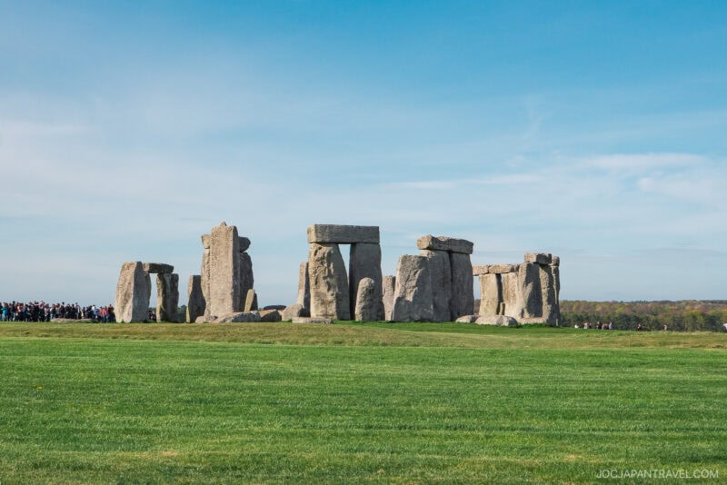 Stonehenge, a prehistoric stone monument of large upright stones in a circle, stands on a grassy field under a clear blue sky with a group of people in the distance, just a short journey from London.