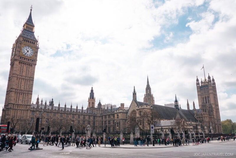 Crowds of people walk in front of the Palace of Westminster and Big Ben in London on a cloudy day, with the iconic clock tower visible on the left and London&rsquo;s stunning gothic architecture dominating the scene.