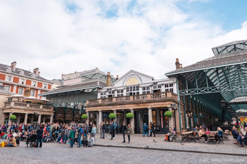 A lively scene at Covent Garden in London shows crowds of people sitting and walking in the cobblestone square, with historic market buildings and outdoor cafes capturing the vibrant spirit of London under a partly cloudy sky.