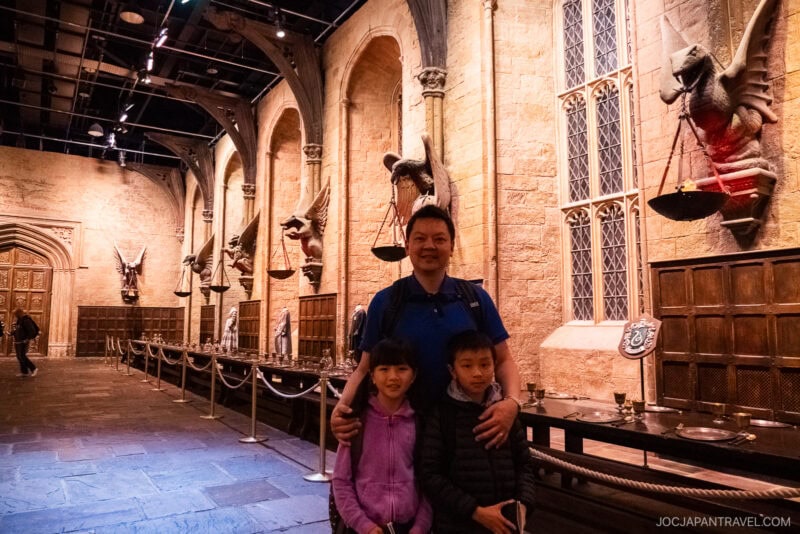 A man stands with two children inside a large, ornate hall in London with high ceilings, long dining tables, stone walls, and statues of mythical creatures. The setting resembles the Hogwarts Great Hall from Harry Potter.