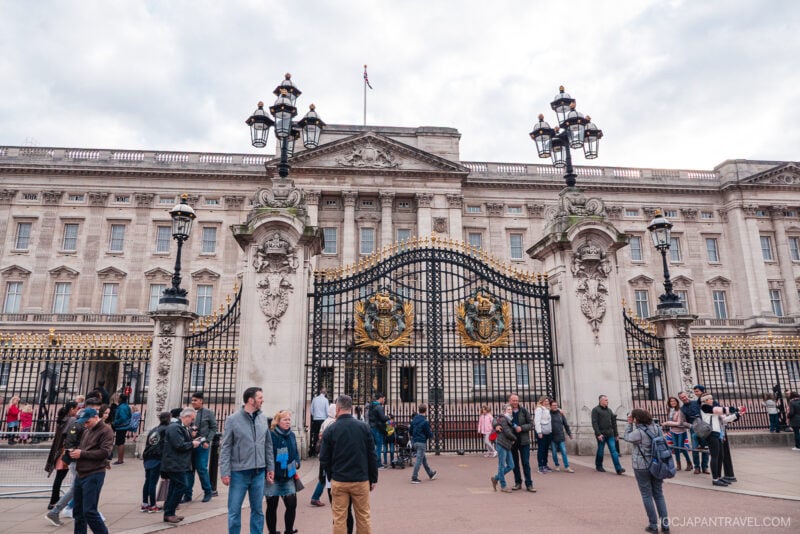 Crowds of people gather in front of the ornate gates of Buckingham Palace in London, with the grand building and classic lampposts visible beneath a cloudy London sky.