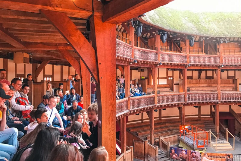 Audience members sit on wooden benches inside a round, open-air London theater with timber beams and a thatched roof, evoking a historical or Shakespearean setting. Some people look towards the mostly empty stage area.