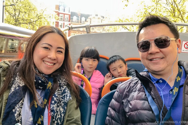 A smiling family of four sits on the upper deck of a London sightseeing bus, wearing audio tour lanyards. Two children sit behind two adults on a sunny day, with buildings and trees visible in the background.