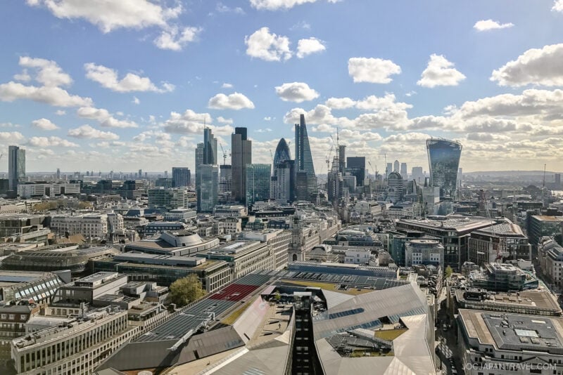 A panoramic view of London’s city skyline featuring modern skyscrapers, including The Gherkin and The Walkie Talkie, under a partly cloudy blue sky with sunlight brightening the iconic London buildings.