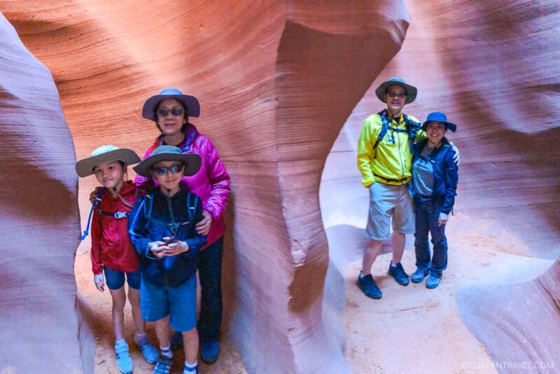 A group of five people wearing hats and outdoor gear stand inside the winding, narrow Lower Antelope Canyon, smiling at the camera as sunlight filters in, highlighting the textured sandstone walls.