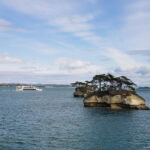 A small rocky island with pine trees sits in blue water under a partly cloudy sky. In the distance, a white sightseeing boat sails across Matsushima Bay.